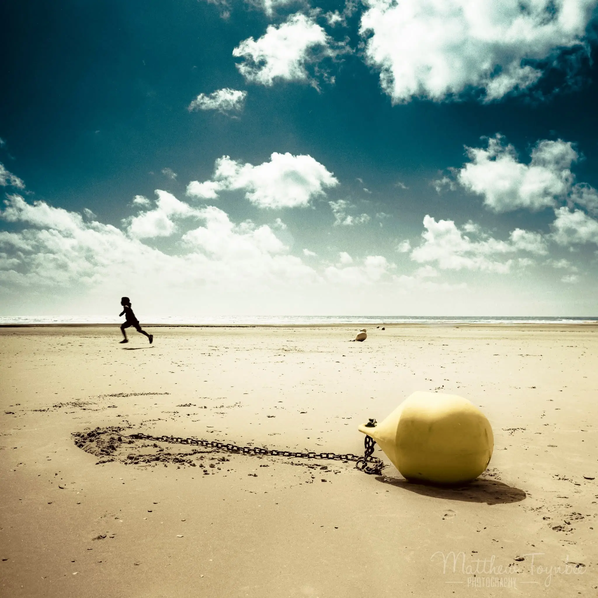 A child running along a wide sandy beach under a blue sky, with a yellow mooring buoy in the foreground